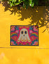 A rectangular polyester doormat with a vibrant, psychedelic design on a hot pink background. In the center is a cute, cartoonish ghost in off-white with yellow and orange outlines; it has large, dark, expressive eyes. The ghost is surrounded by a maximalist pattern of 70s-style floral motifs and puffy clouds in shades of teal, blue, orange, and light pink. Two small, colorful rainbows arc over the ghost's shoulders. The doormat has a durable, textured surface and is framed by a sleek black trim.